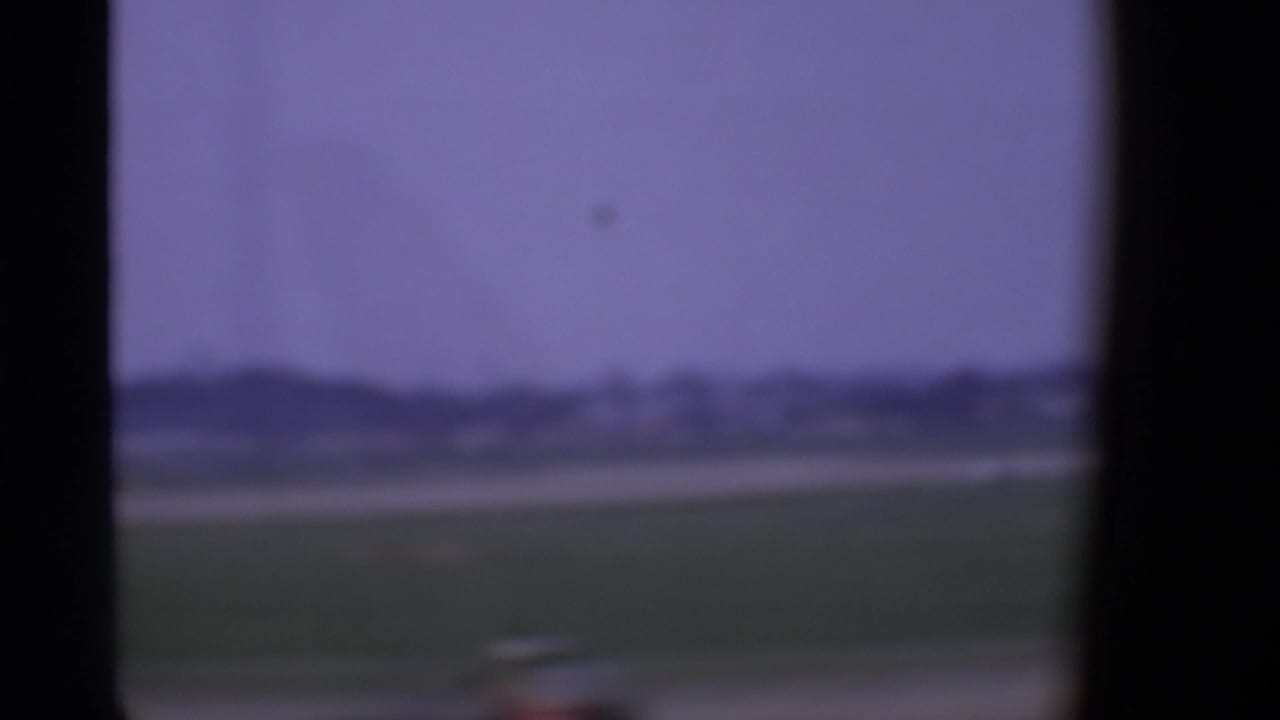 Soft 1969 London airport view through an aircraft window, with grass, distant low buildings, and hazy sky.