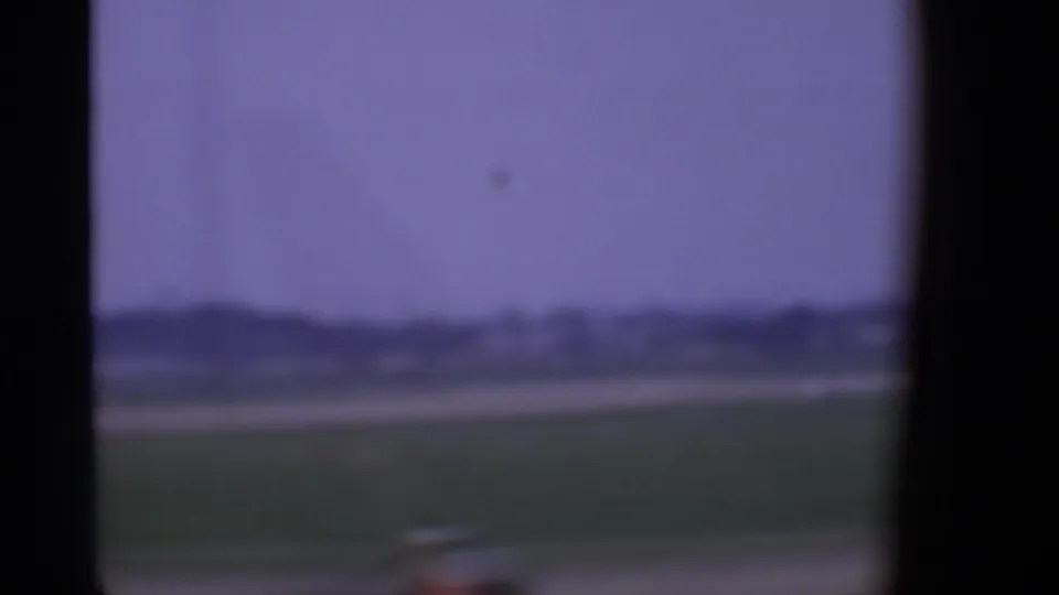 Soft 1969 London airport view through an aircraft window, with grass, distant low buildings, and hazy sky.