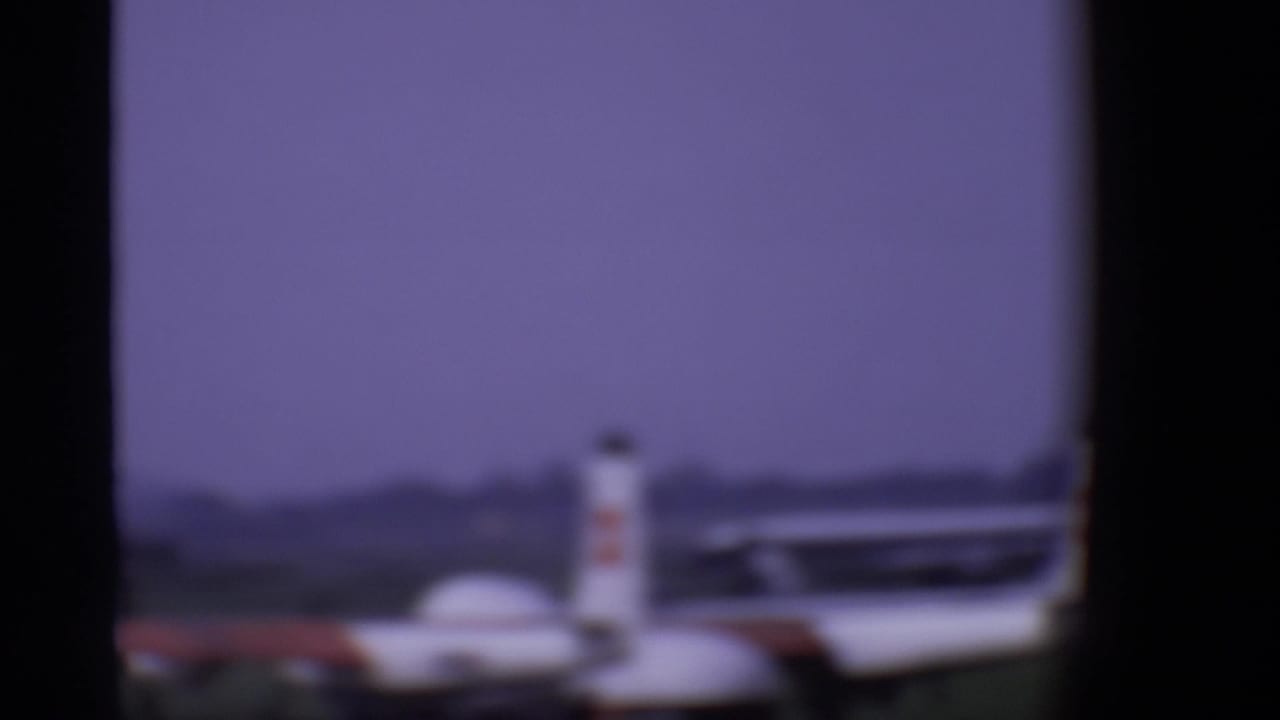 A blurred small aircraft appears on the London airport tarmac beyond dark airplane window framing in 1969 footage.