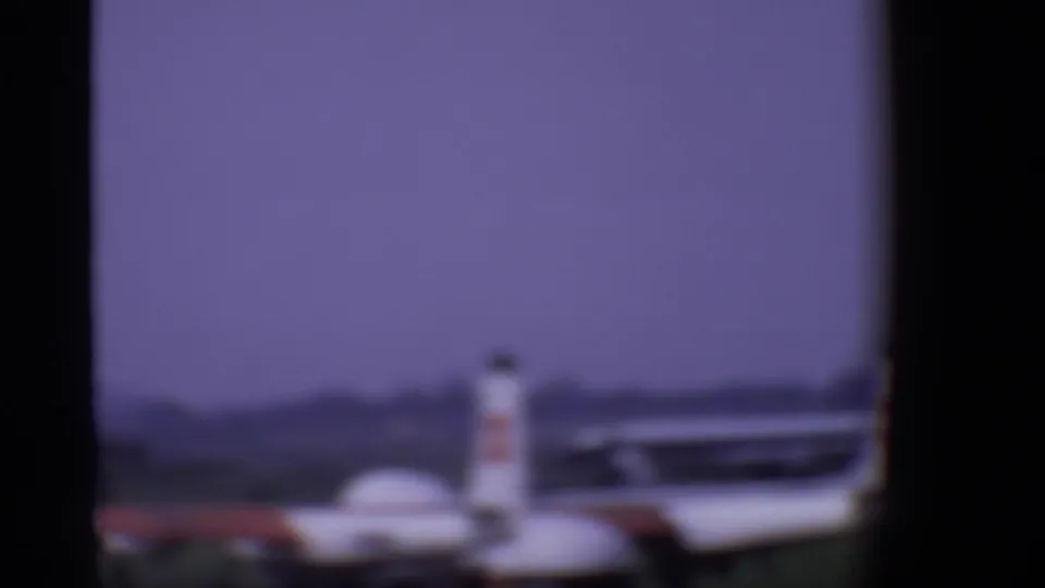 A blurred small aircraft appears on the London airport tarmac beyond dark airplane window framing in 1969 footage.