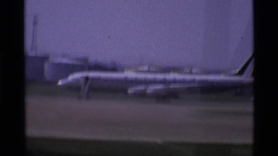 A parked airplane is visible on the London airport apron, seen in soft 8mm focus with low buildings behind it.