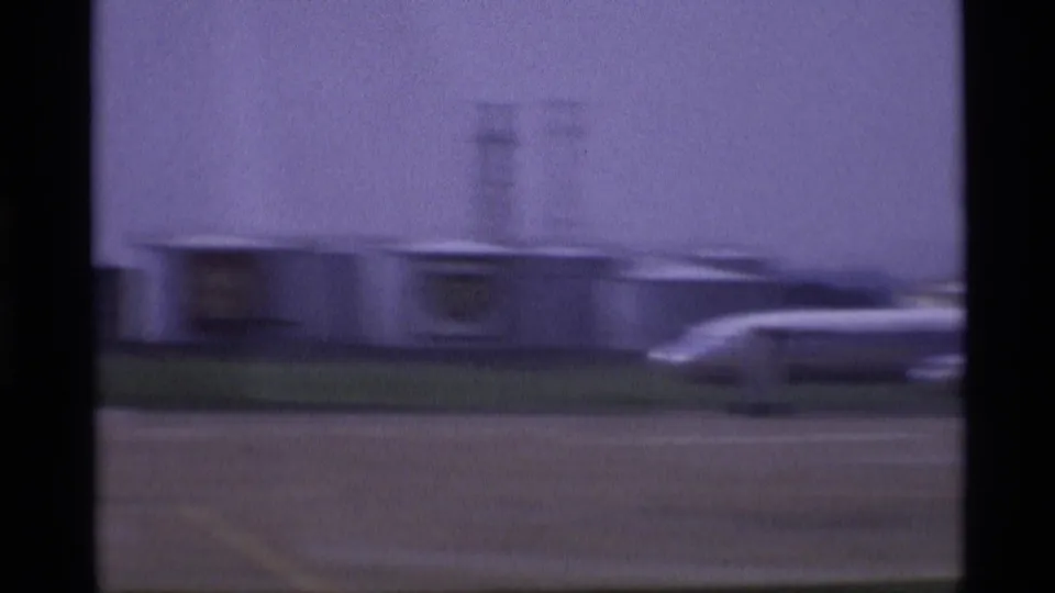 Low airport structures and a partial aircraft appear across the London tarmac in purple-tinted 1969 home-movie footage.
