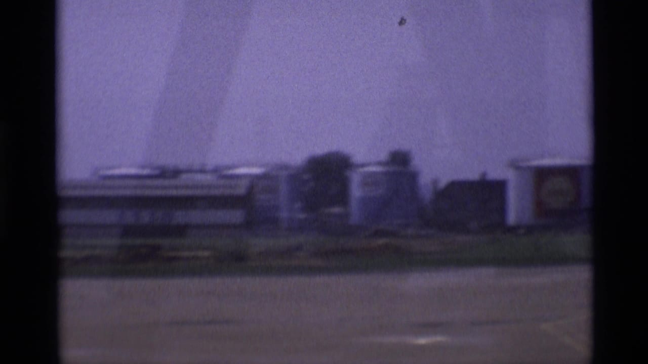 1969 London home-movie frame showing low industrial structures and cylindrical tanks beyond pavement, partly obscured by dark vertical window edges.