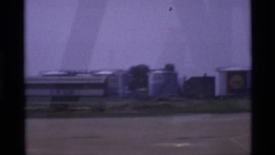 1969 London home-movie frame showing low industrial structures and cylindrical tanks beyond pavement, partly obscured by dark vertical window edges.