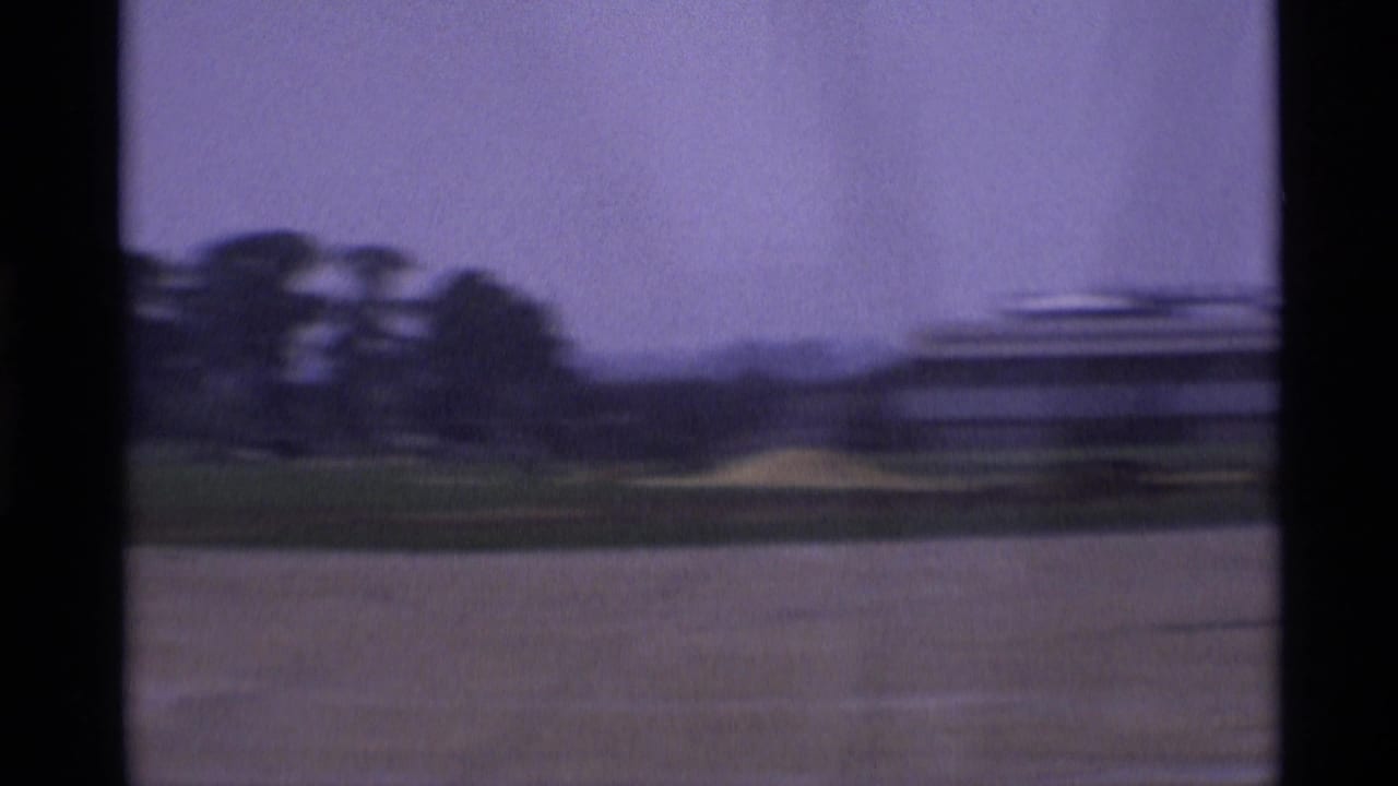 1969 London suburban outskirts with open grass, distant trees, and blurred foreground, seen through dark vertical framing during a pan.