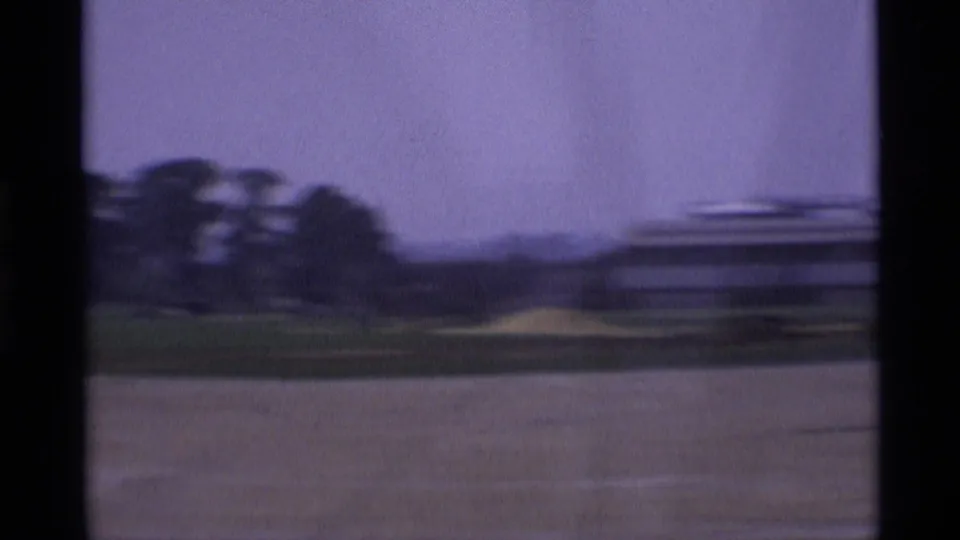 1969 London suburban outskirts with open grass, distant trees, and blurred foreground, seen through dark vertical framing during a pan.