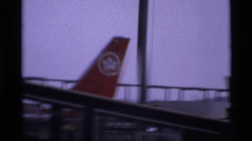 1969 London frame showing a red aircraft tail with a visible logo, seen behind dark structural bars or window framing near airport-edge scenery.