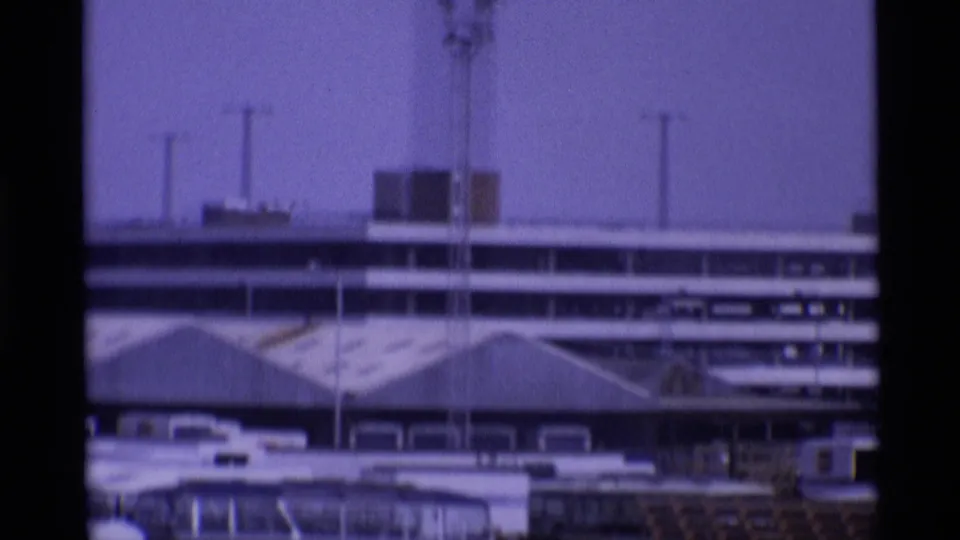 1969 London airport frame showing terminal rooflines, an antenna mast, parked vehicles, and apron-side buildings.