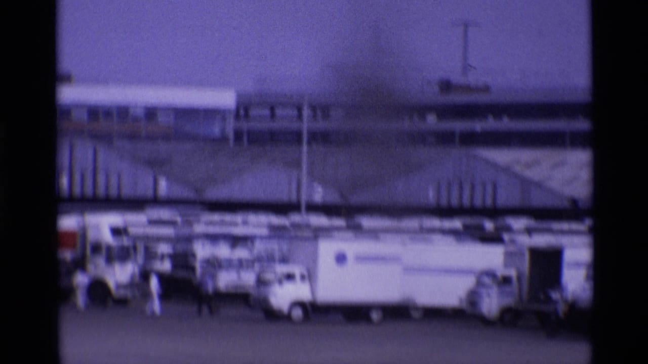 Airport apron with service vehicles, ground crew, and terminal buildings behind a stationary aircraft area.