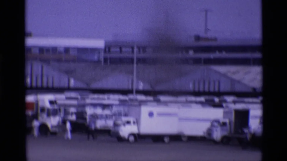 1969 London airport tarmac view with service trucks, small figures, and terminal structures seen through soft purple 8mm film.
