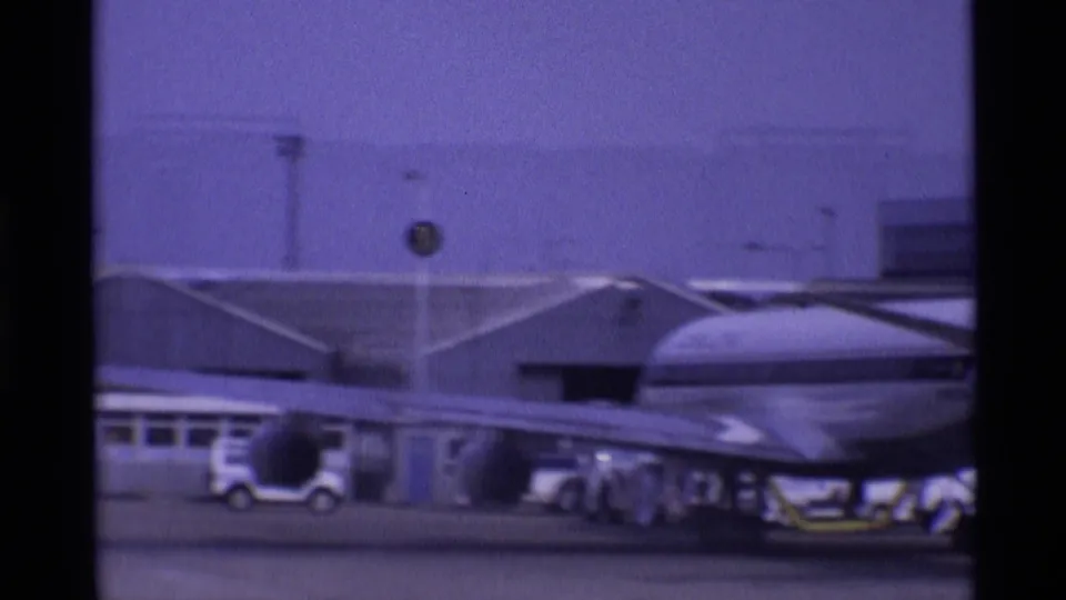 1969 Heathrow-area tarmac view showing an aircraft wing, service activity, and low airport buildings on purple-shifted 8mm film.