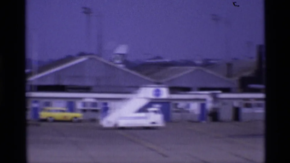 London airport apron in 1969 with jet stairs, service vehicles, terminal buildings, and faint smoke or haze beyond the roofline.