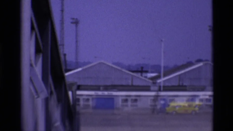 1969 London airport buildings viewed through dark framing, with blue doors and a yellow service vehicle or taxi near the terminal.