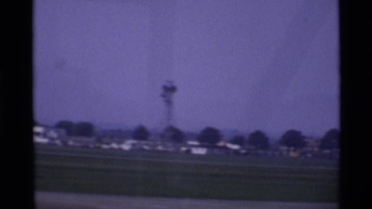 1969 London airport airfield view with grass, distant low buildings, and a tower-like structure beyond the tarmac.