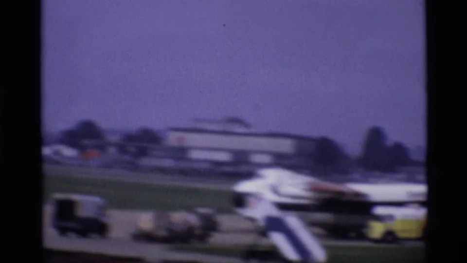 1969 London airport apron with mobile aircraft stairs, small ground vehicles, and a blurred aircraft form in the background.