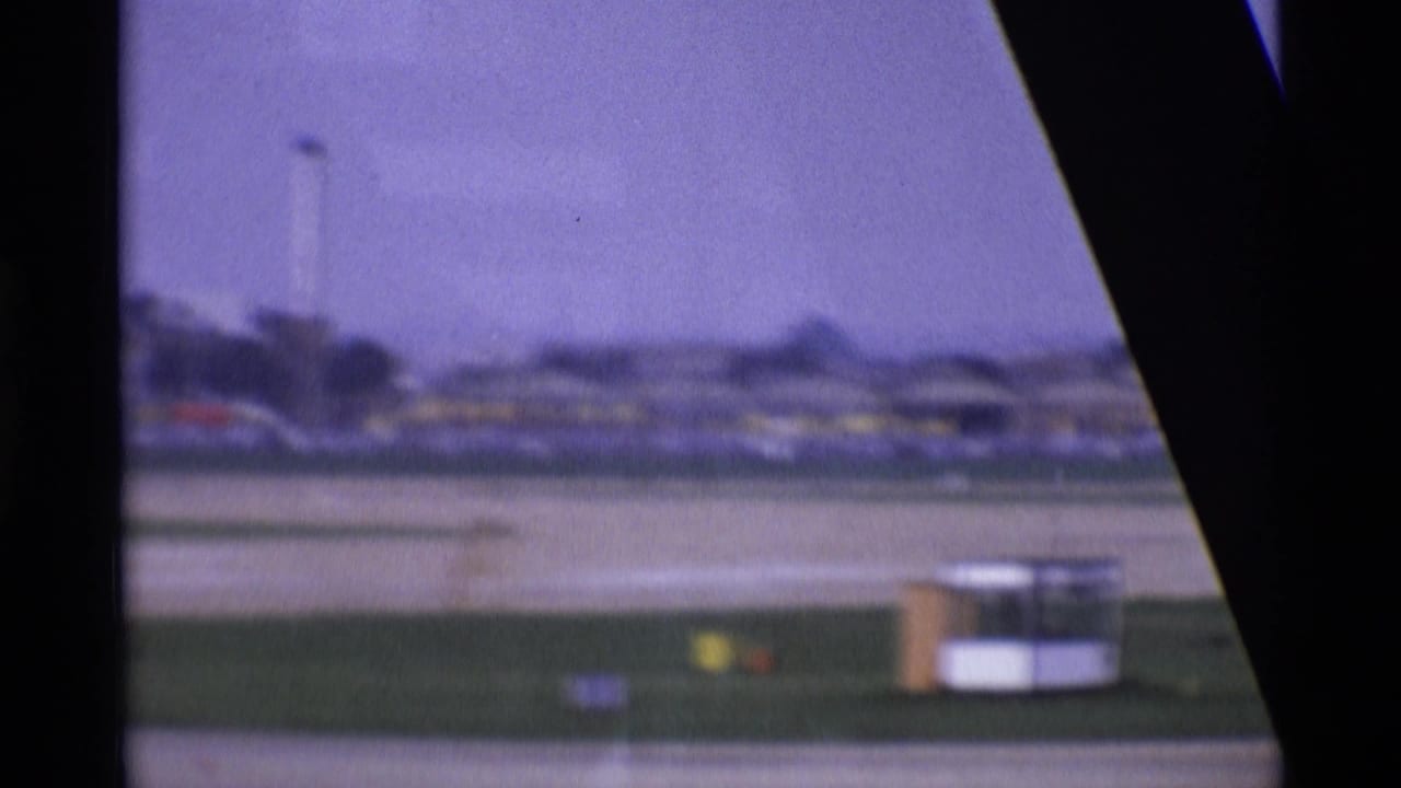 1969 London airport runway seen through an aircraft window, with terminal structures and a soft purple 8mm film cast.