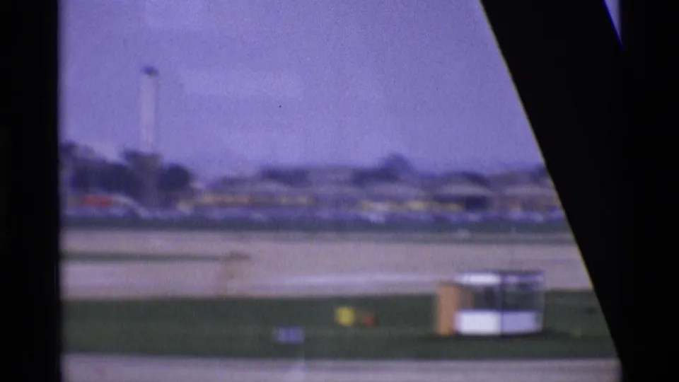 1969 London airport runway seen through an aircraft window, with terminal structures and a soft purple 8mm film cast.