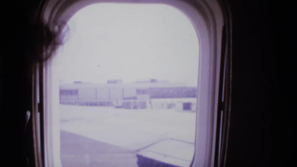 1969 London airplane cabin view through a rounded window toward terminal buildings and tarmac.