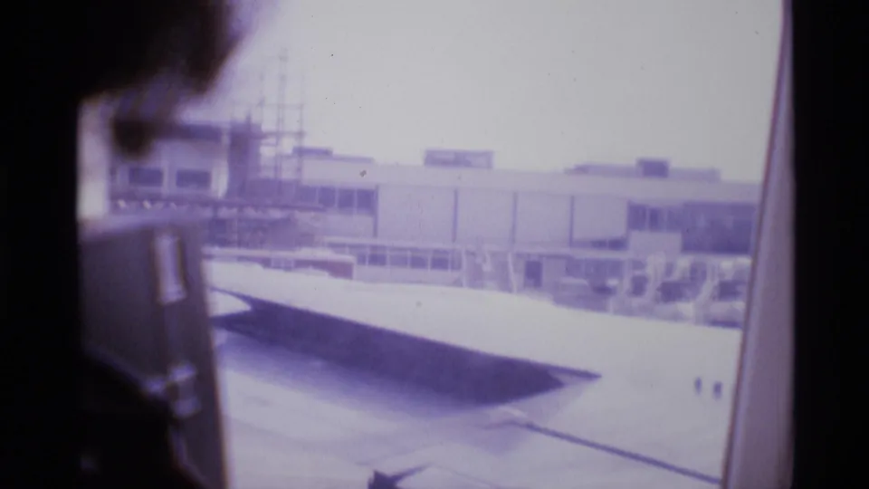 Passenger silhouette beside an airplane window with London airport buildings and wing visible beyond.