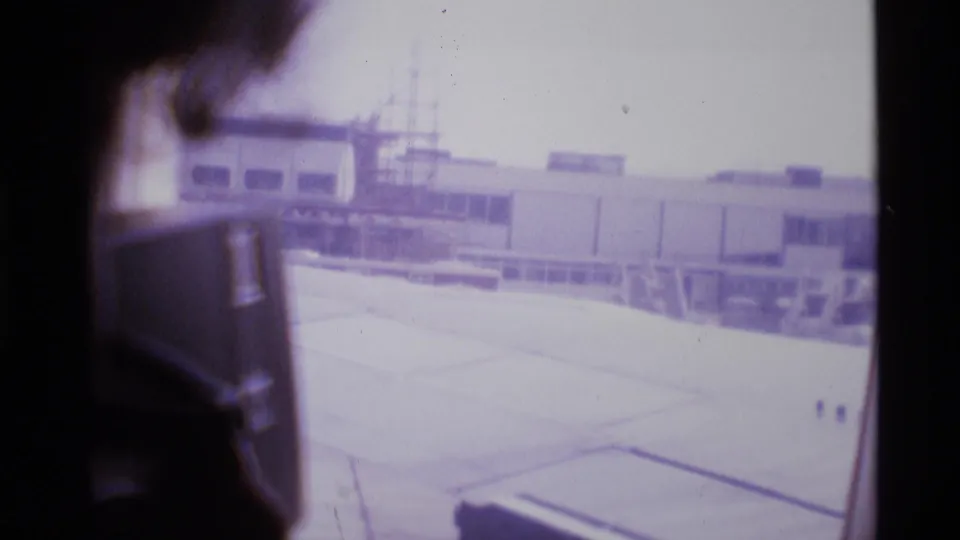 1969 airplane window view with a passenger silhouette, wing, tarmac, and London airport terminal structures outside.