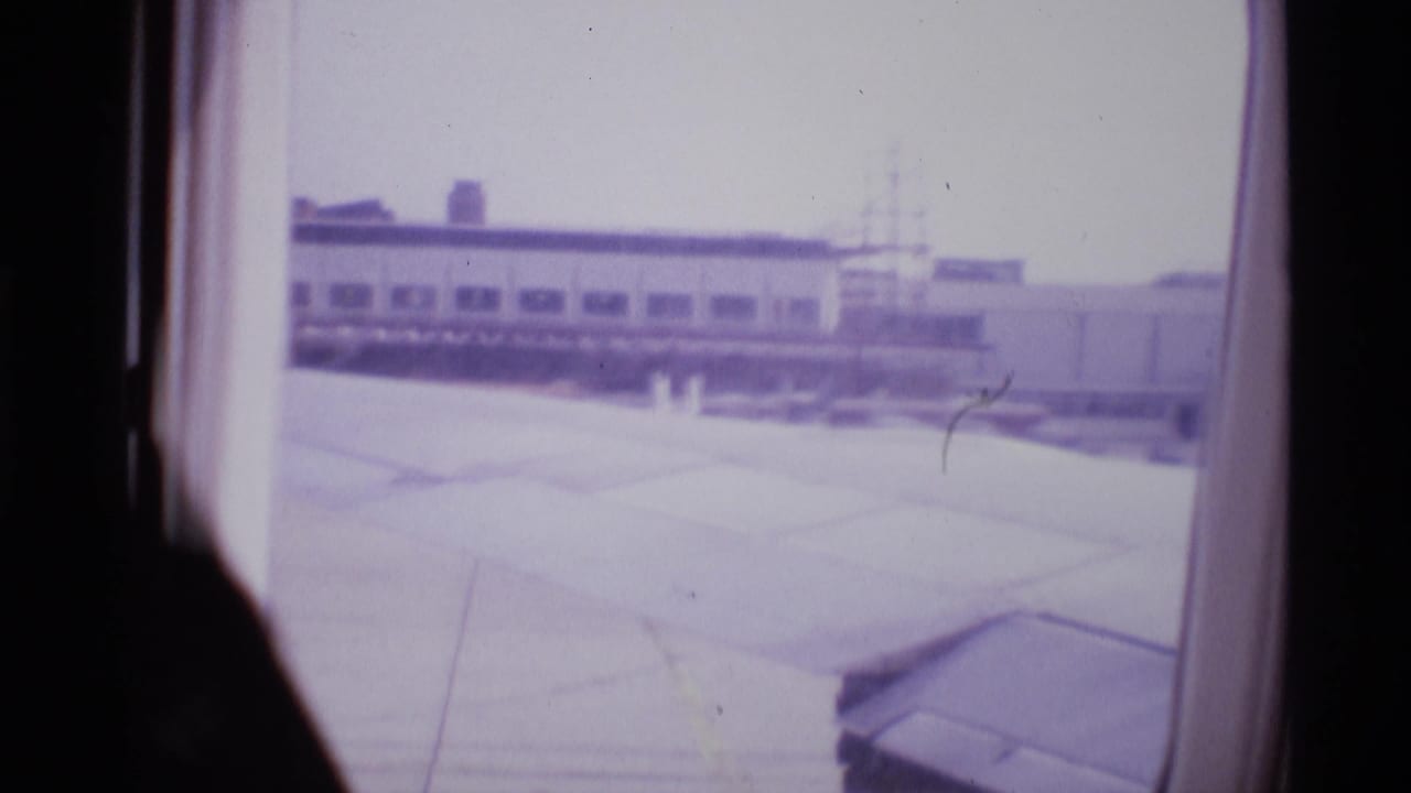 1969 London airport departure view through an airplane window, with the wing foreground and low terminal buildings beyond.