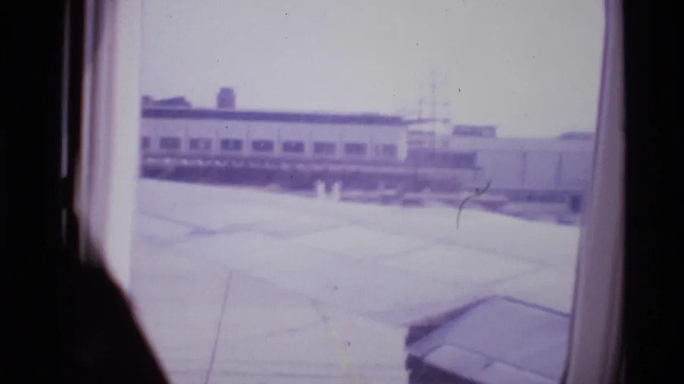 1969 London airport departure view through an airplane window, with the wing foreground and low terminal buildings beyond.