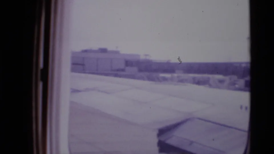 Soft 8mm view from a departing plane at a London airport, showing the wing, apron, and distant terminal structures.