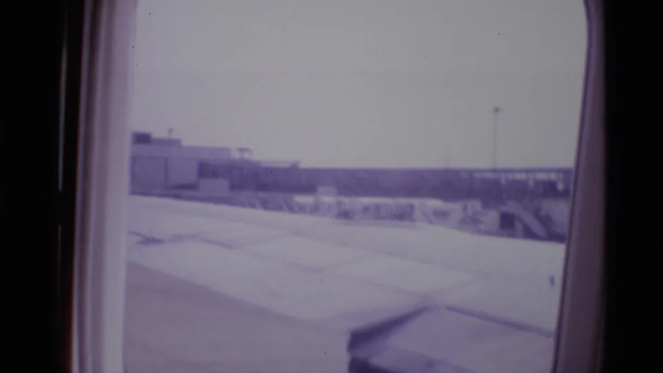 Passenger-shot 1969 London airport view with wing foreground, pale tarmac, and low buildings beyond the cabin window.