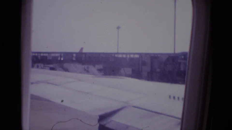 London airport scene from a 1969 airplane window, with the wing, apron surface, and distant airport structures under a pale sky.