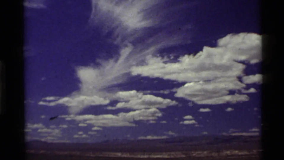 1972 8mm footage holds on a blue sky with layered white clouds over distant hills and open terrain.