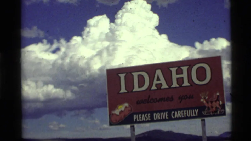 1972 roadside travel footage shows an Idaho welcome sign reading "Please drive carefully" below towering white clouds.