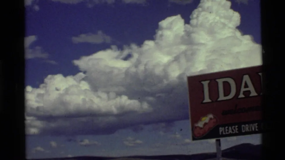 Moving 8mm frame with the Idaho welcome sign partly cropped at right and expansive cumulus clouds over low hills.