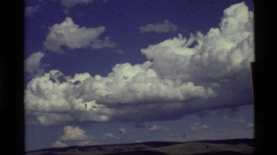 1972 8mm travel frame showing blue sky, layered white clouds, and a low western horizon after the roadside sign has passed.