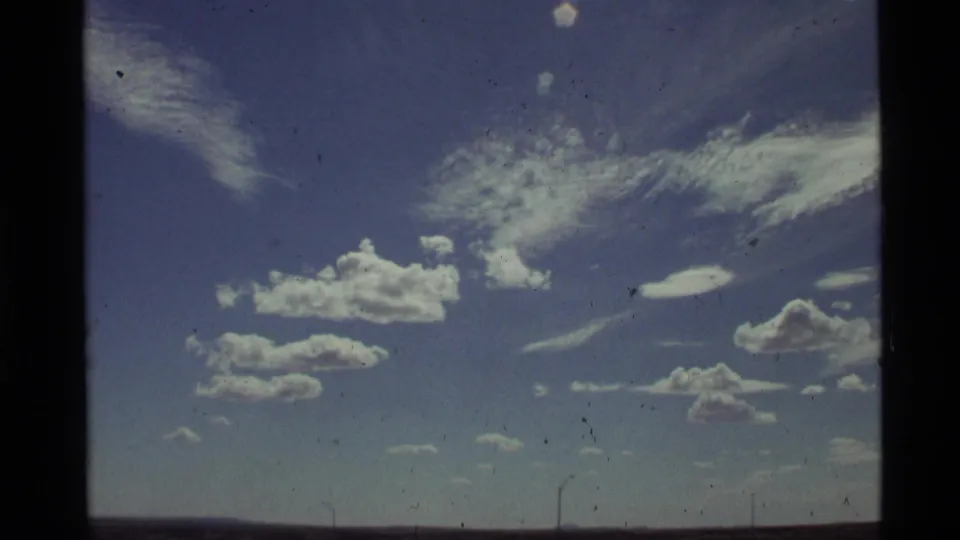 1972 Holland Lake, Montana home-movie frame showing scattered clouds, thin cirrus, and faint roadside poles near the horizon.