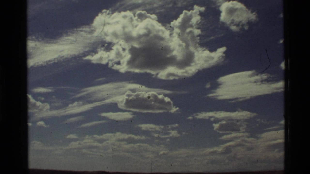 1972 Holland Lake, Montana 8mm frame showing bright cumulus clouds, wispy layers, blue sky, and a faint distant horizon.