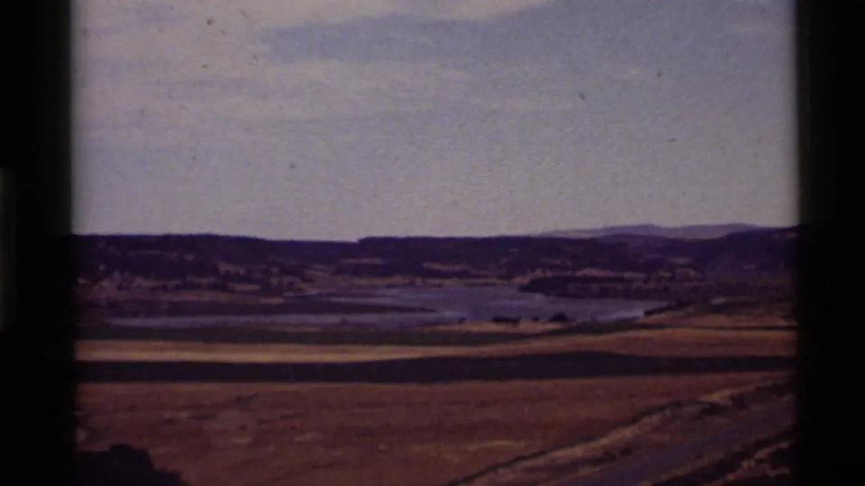 Wide 1972 Holland Lake landscape with dry foreground fields, calm water, distant hills, and a hazy sky in faded 8mm color.