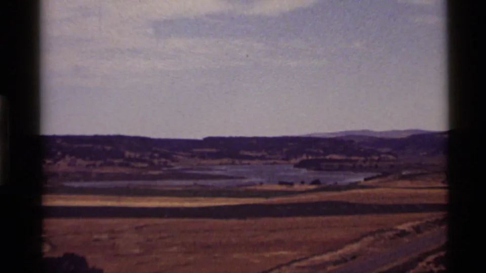 A 1972 8mm view of Holland Lake, Montana, with sunlit dry fields, calm water, distant ridges, and visible home-movie grain.