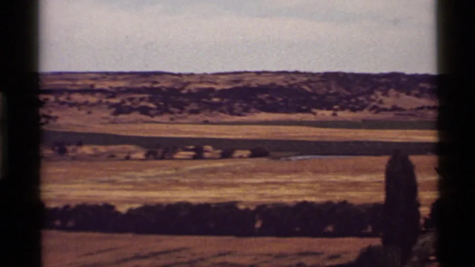 1972 home-movie panorama near Holland Lake, Montana, with golden fields, low shrubs, distant hills, and a pale sky.