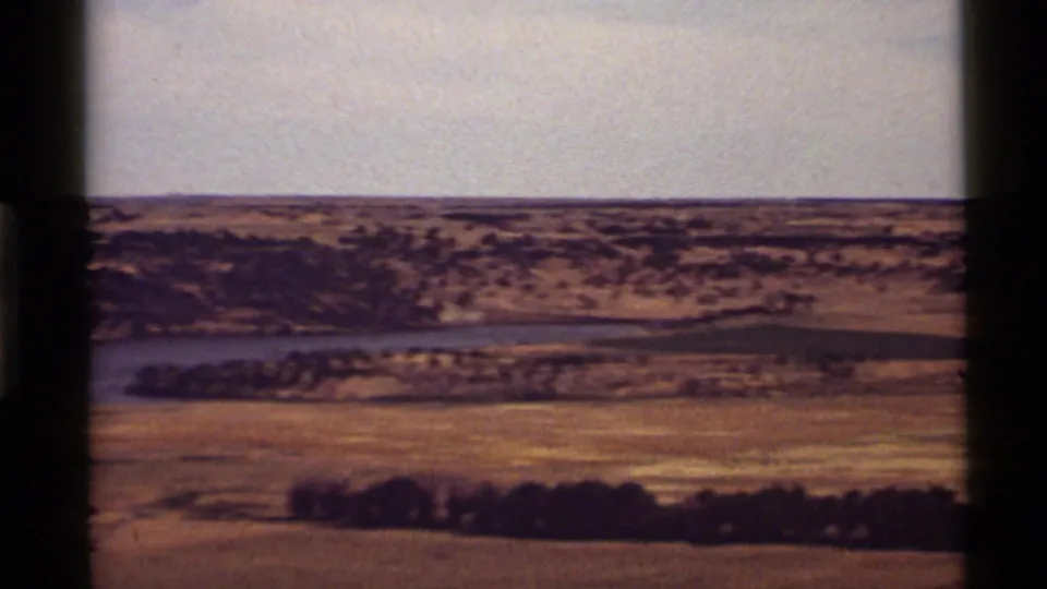 1972 8mm Montana landscape with a river or lake band cutting across dry open land beneath a distant ridge.