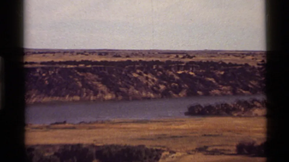 1972 Holland Lake area view showing broad water, dry shoreline, scrub-covered slope, and pale sky in warm 8mm color.