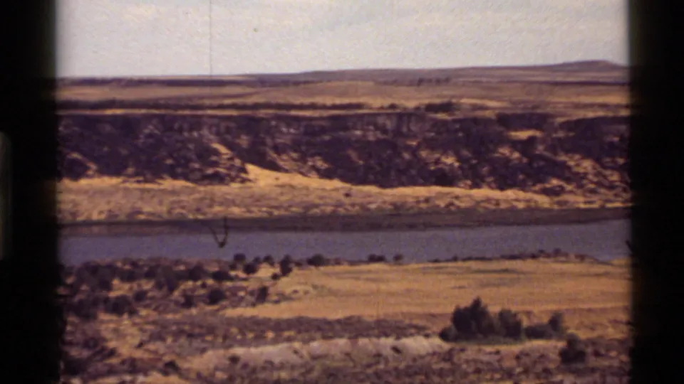 8mm frame of the Montana landscape with a dry hillside, blue water below, scattered shrubs, pale sky, and light film wear.