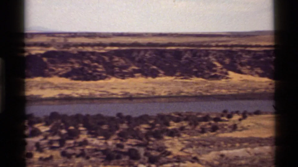 Final frame from the 1972 Montana pan showing blue water, a dark eroded bank, brush in the foreground, and open plains beyond.