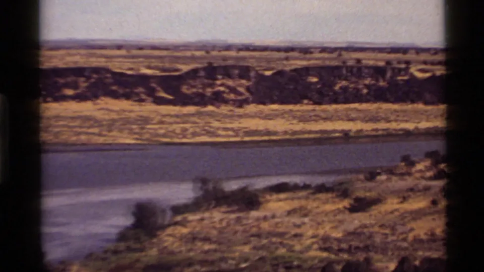 1972 8mm frame from Holland Lake, Montana, with a curving river bend, dry grassy banks, shrubs, and a broad open horizon.