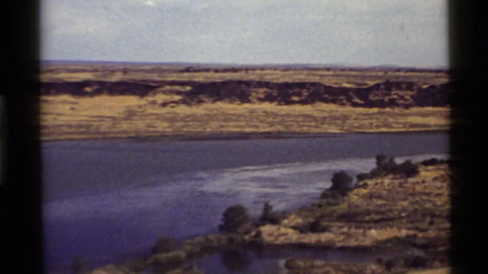 1972 Holland Lake, Montana 8mm frame showing a river bend bordered by scrubby shoreline, dry terrain, and low distant ridges.