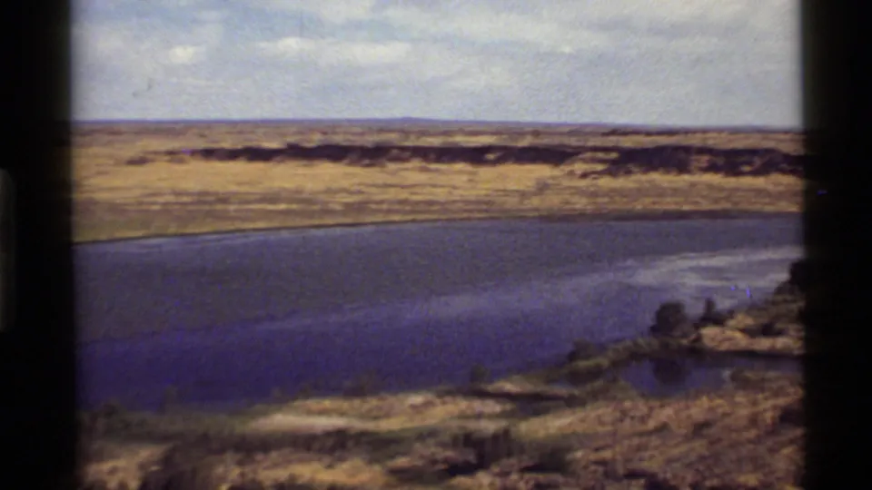 1972 8mm Montana river view with blue-green water, dry open banks, sparse trees, and distant low ridges under a pale sky.