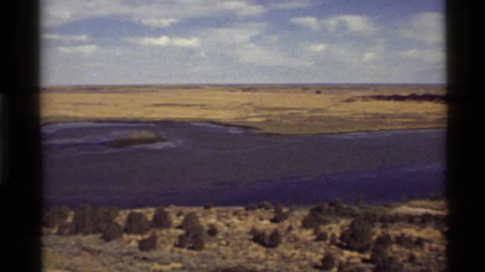 Home-movie frame from 1972 near Holland Lake, Montana, with shoreline, wheat-colored fields, brush, and a low horizon.
