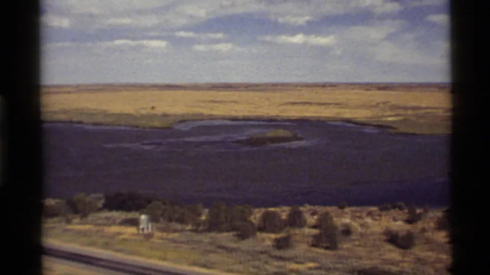 1972 8mm view near Holland Lake, Montana, with water, open fields, trees, and a small distant structure or object.