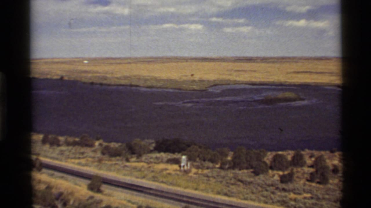 1972 Montana home-movie frame showing dark water, open dry terrain, a roadside strip, and cloudy sky from a moving viewpoint.