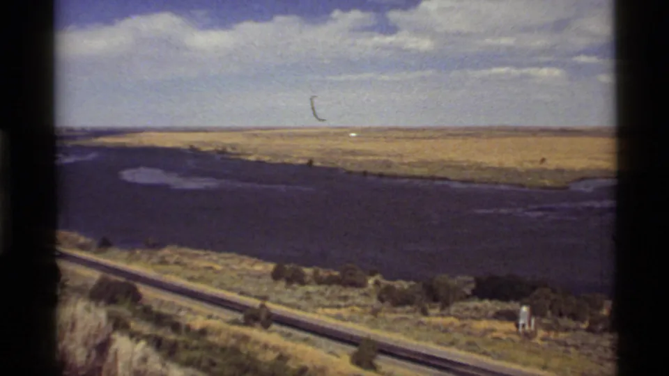 1972 Montana 8mm frame with a highway running beside dark water, low brush, golden ground, and a partly cloudy horizon.