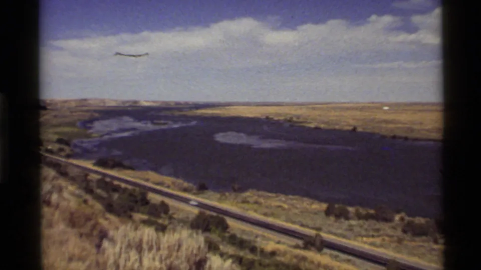 1972 Montana travel frame showing a curving waterway near a highway, scattered trees, dry slopes, and a bright clouded sky.